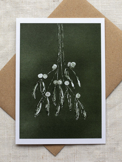 Botanical Christmas card with mistletoe design on a dark green background, placed on a linen surface with a Kraft paper envelope.