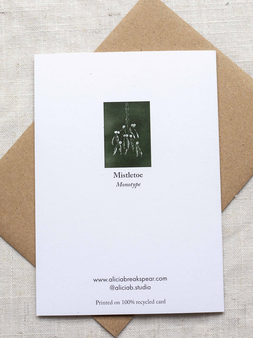 Reverse side of botanical Christmas card with mistletoe design on a dark green background, placed on a linen surface with a Kraft paper envelope.