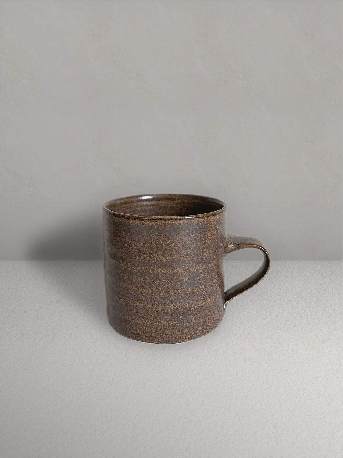 Ceramic mug with low handle in brown glaze on a light taupe surface with a textured taupe wall behind - front view.