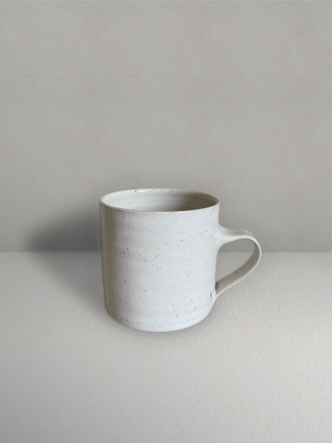 Ceramic mug with low handle in glossy off-white speckled glaze on a light taupe surface with a textured taupe wall behind - front view.