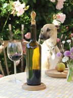 Wine bottle with oak stopper on a table in a garden setting with a glass of wine, flowers and a dog in the background.