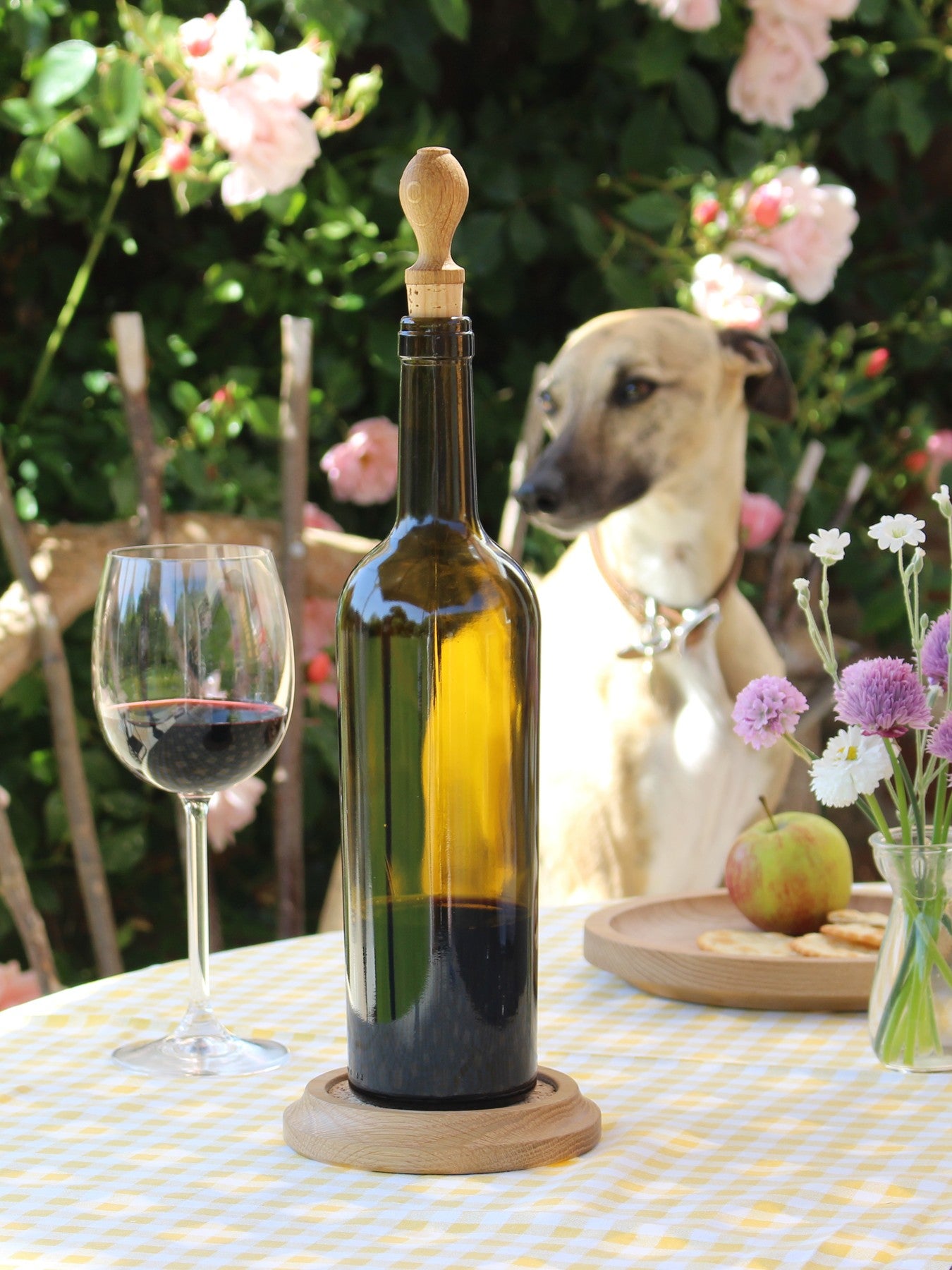 Wine bottle with oak stopper on a table in a garden setting with a glass of wine, flowers and a dog in the background.