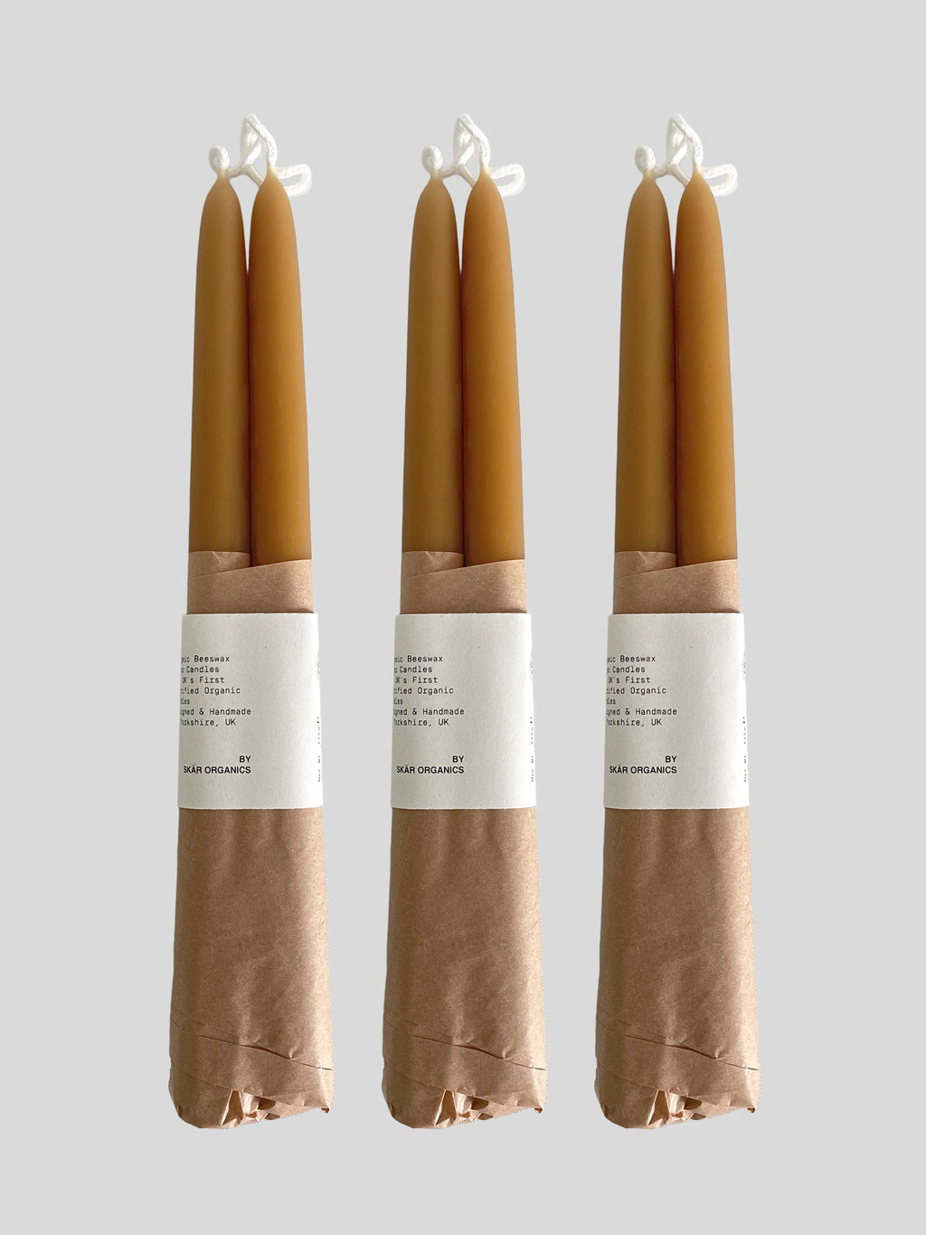 Three pairs of beeswax hand-dipped taper candles in a natural honey colour, wrapped in brown kraft paper with a white paper label, against a solid light neutral background.
