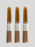 Three pairs of beeswax hand-dipped taper candles in a natural honey colour, wrapped in brown kraft paper with a white paper label, against a solid light neutral background.