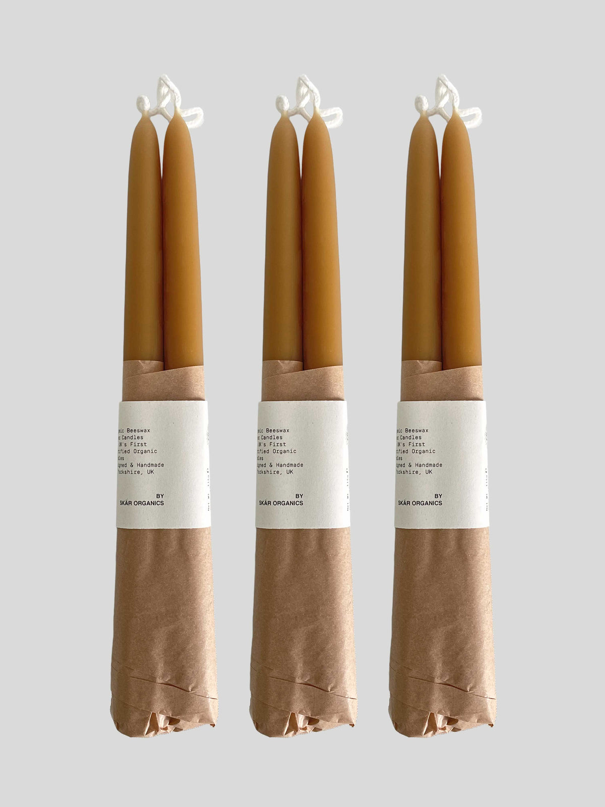 Three pairs of beeswax hand-dipped taper candles in a natural honey colour, wrapped in brown kraft paper with a white paper label, against a solid light neutral background.