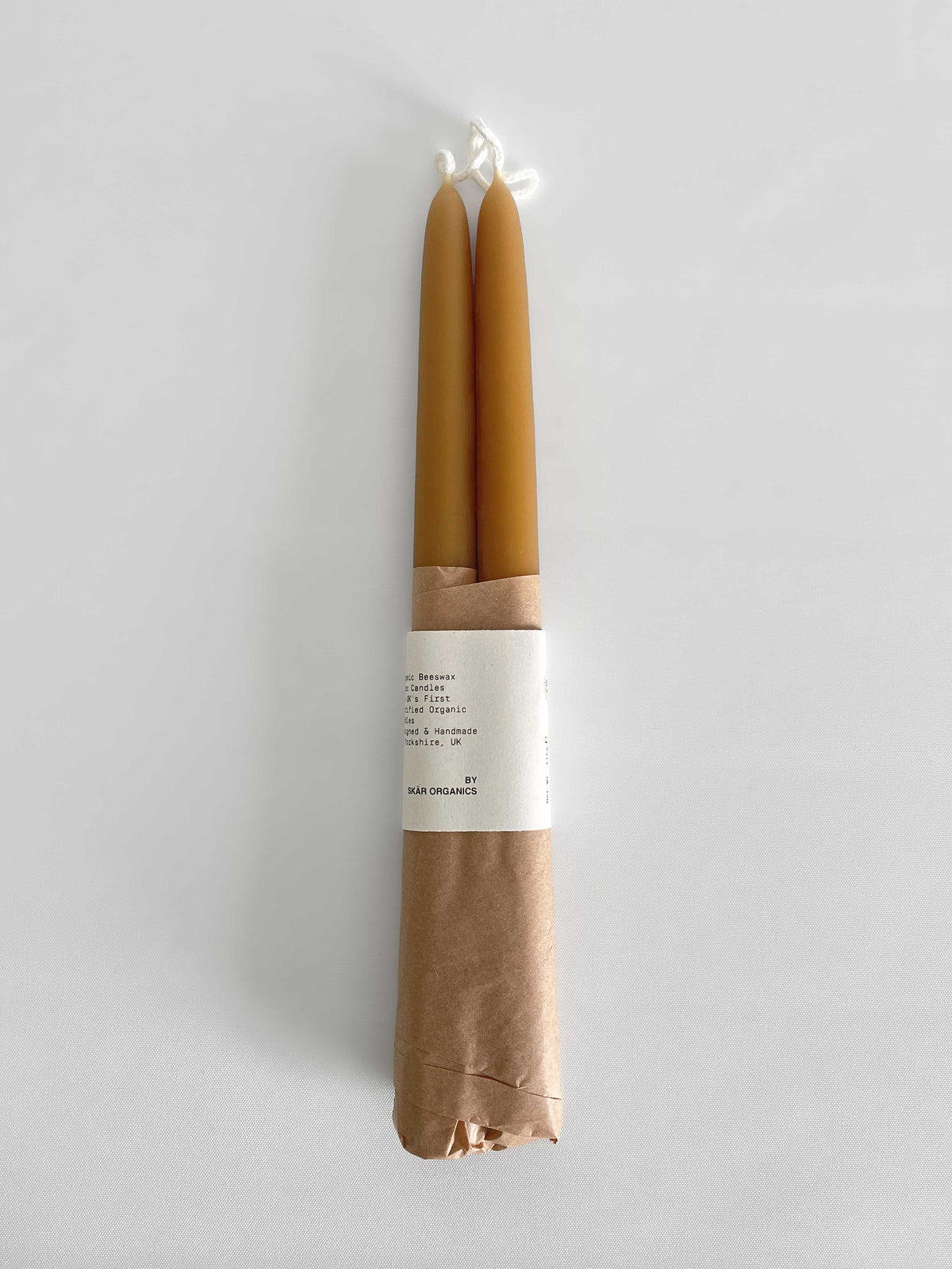 Pair of beeswax hand-dipped taper candles in a natural honey colour, wrapped in brown kraft paper with a white paper label, against a light neutral background.