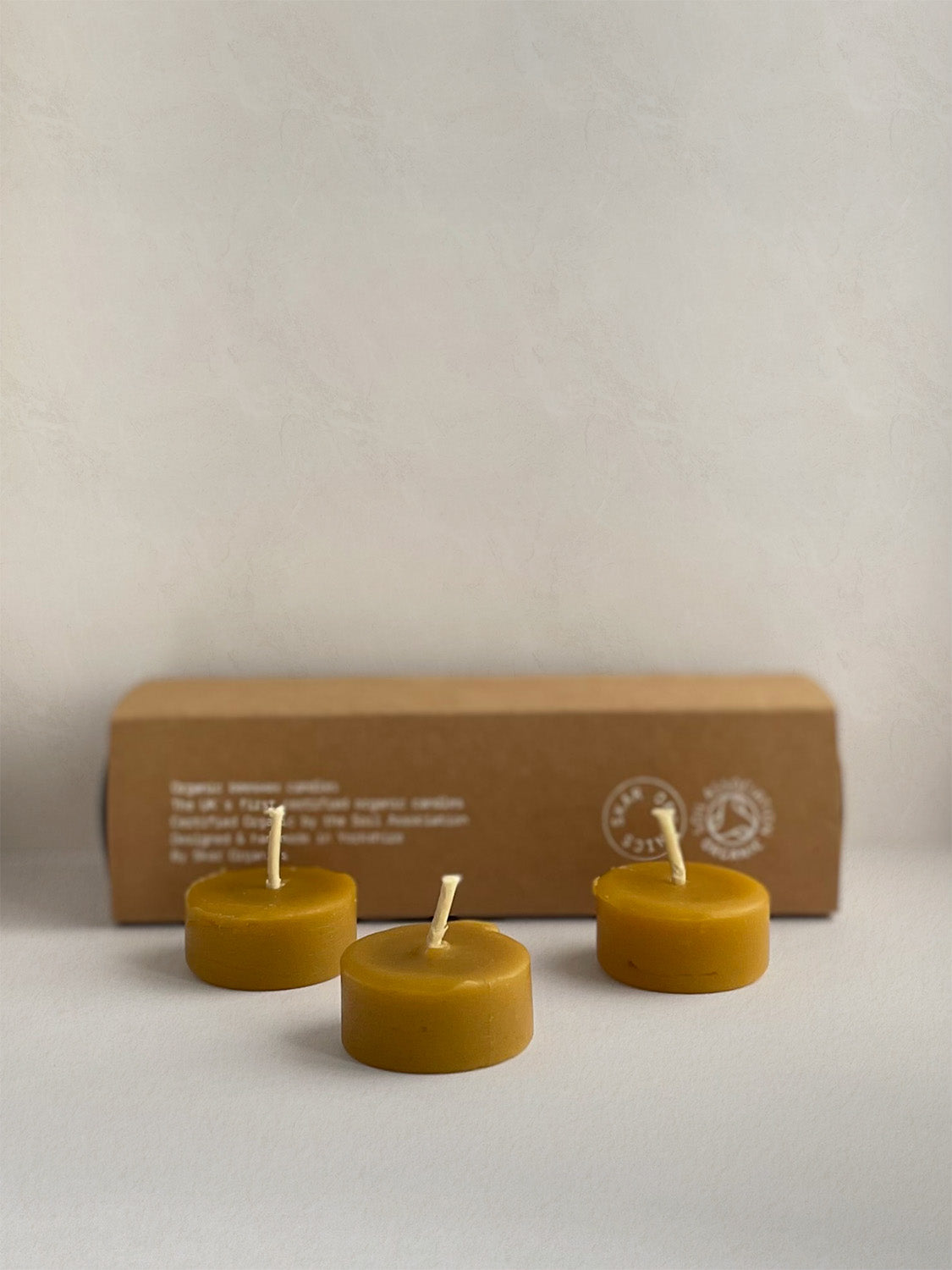 Three organic beeswax tealight candles in front of box on a light surface.