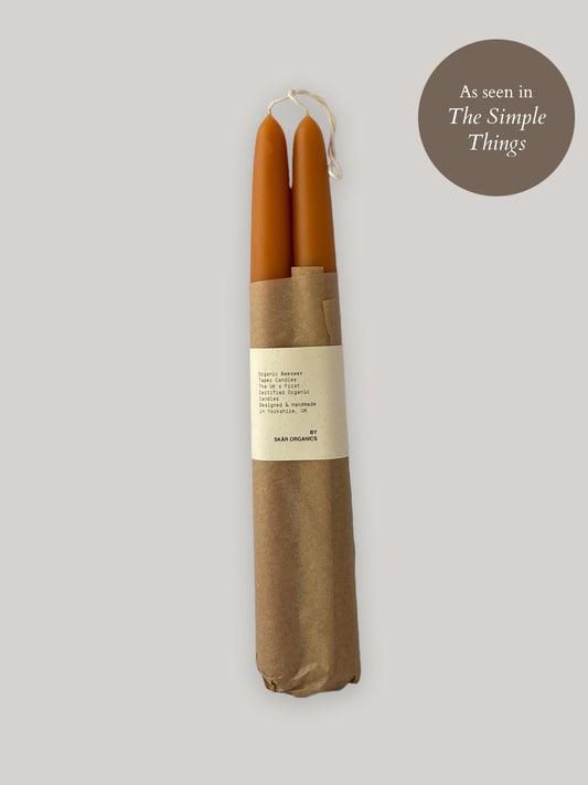 Two orange beeswax taper candles in a brown paper wrap on a neutral background.