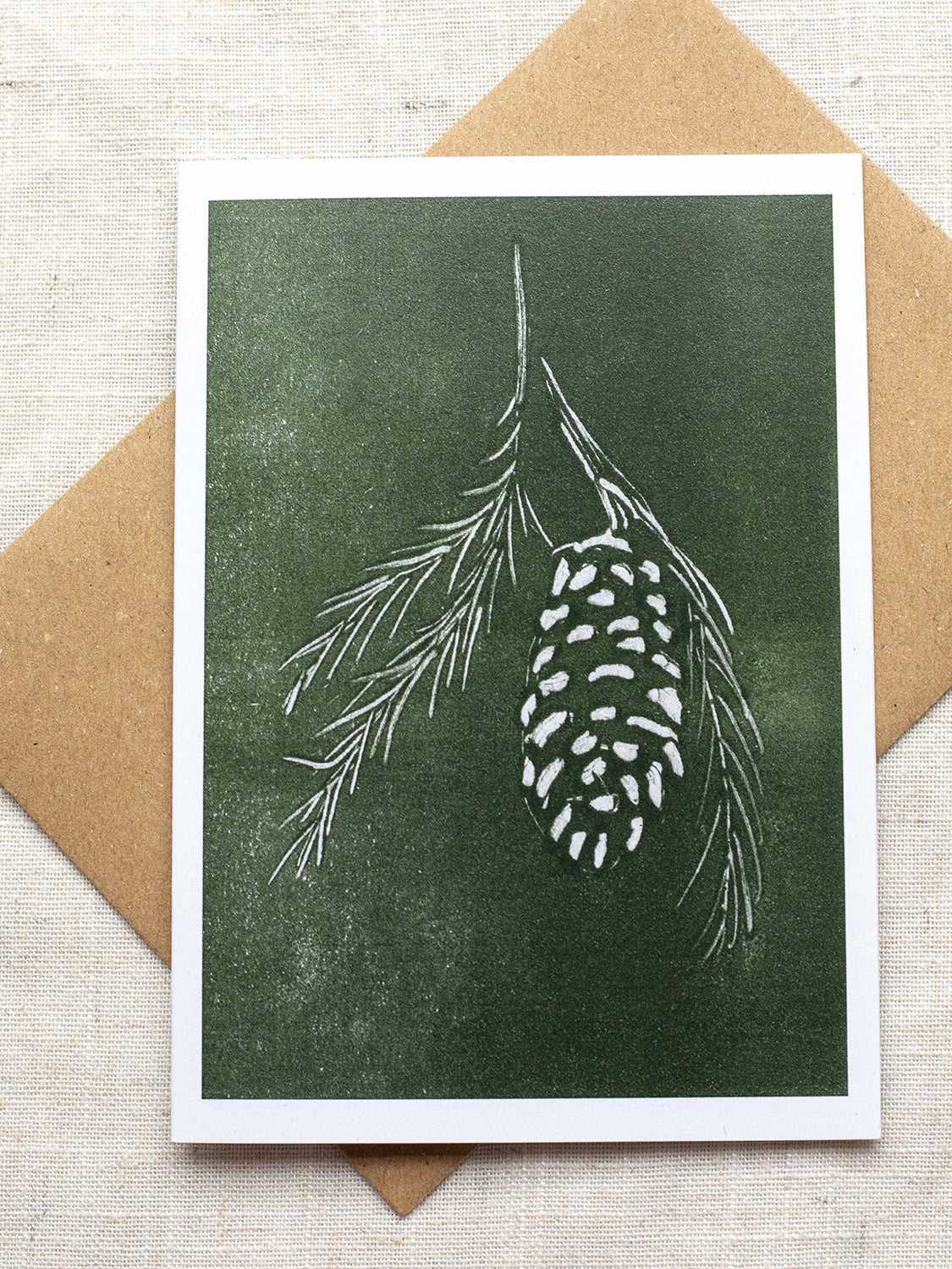 Botanical Christmas card with pine cone and branch design on a green background, placed on a Kraft paper envelope on a linen background.