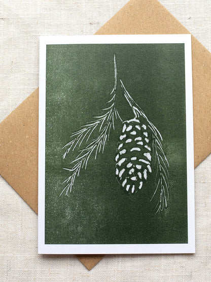 Botanical Christmas card with pine cone and branch design on a green background, placed on a Kraft paper envelope on a linen background.