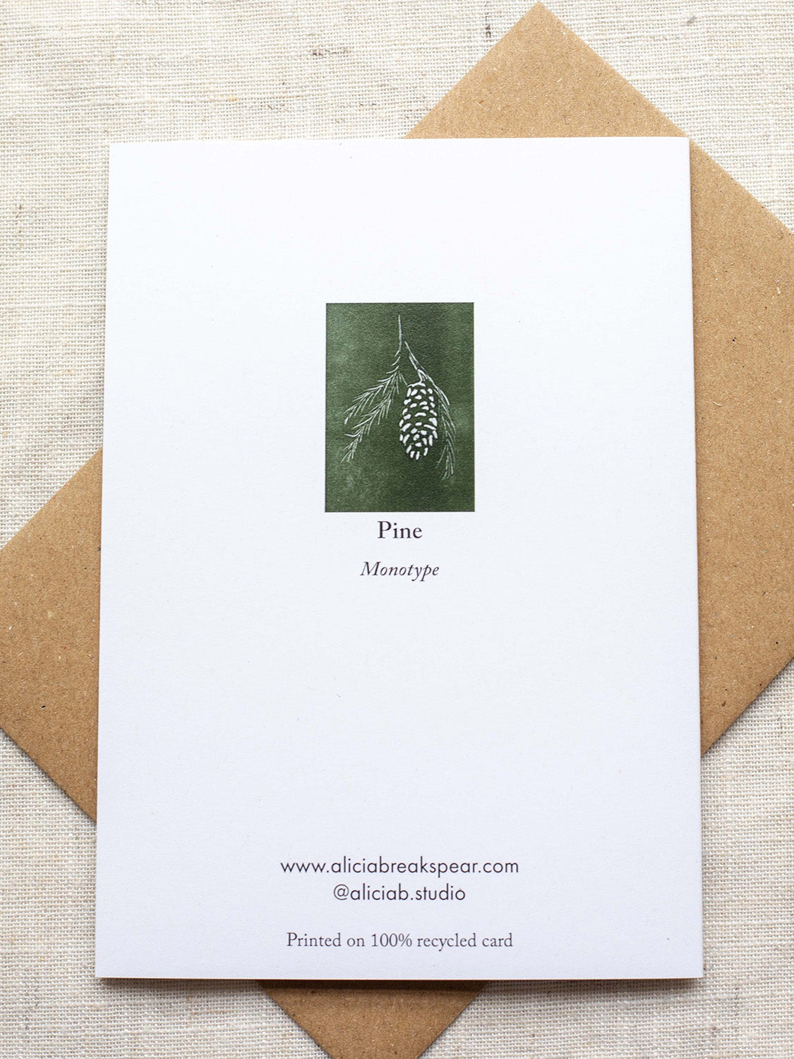 Reverse side of botanical Christmas card with pine cone and branch design on a green background, placed on a Kraft paper envelope on a linen background.