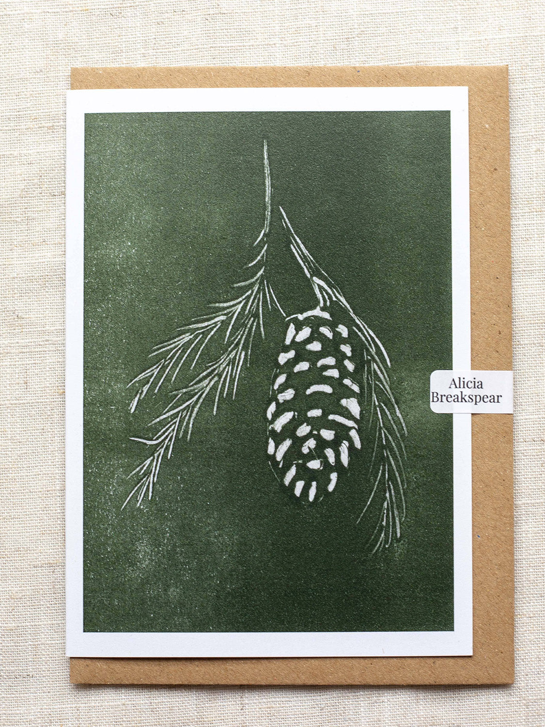 Botanical Christmas card with pine cone and branch design on a green background, with a Kraft paper envelope and 'Alicia Breakspear' sticker seal.