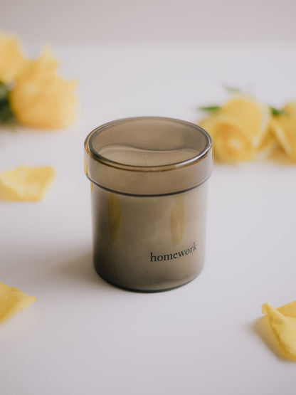 Candle in smoked glass jar labelled 'homework' on a light surface with yellow flowers.