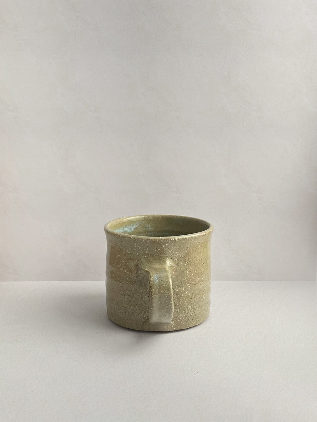 A rustic light green handmade ceramic espresso mug with a handle, on a neutral background - back view.