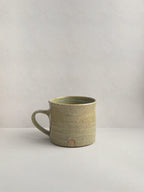 A rustic light green handmade ceramic espresso mug with a handle, on a neutral background - right side view.