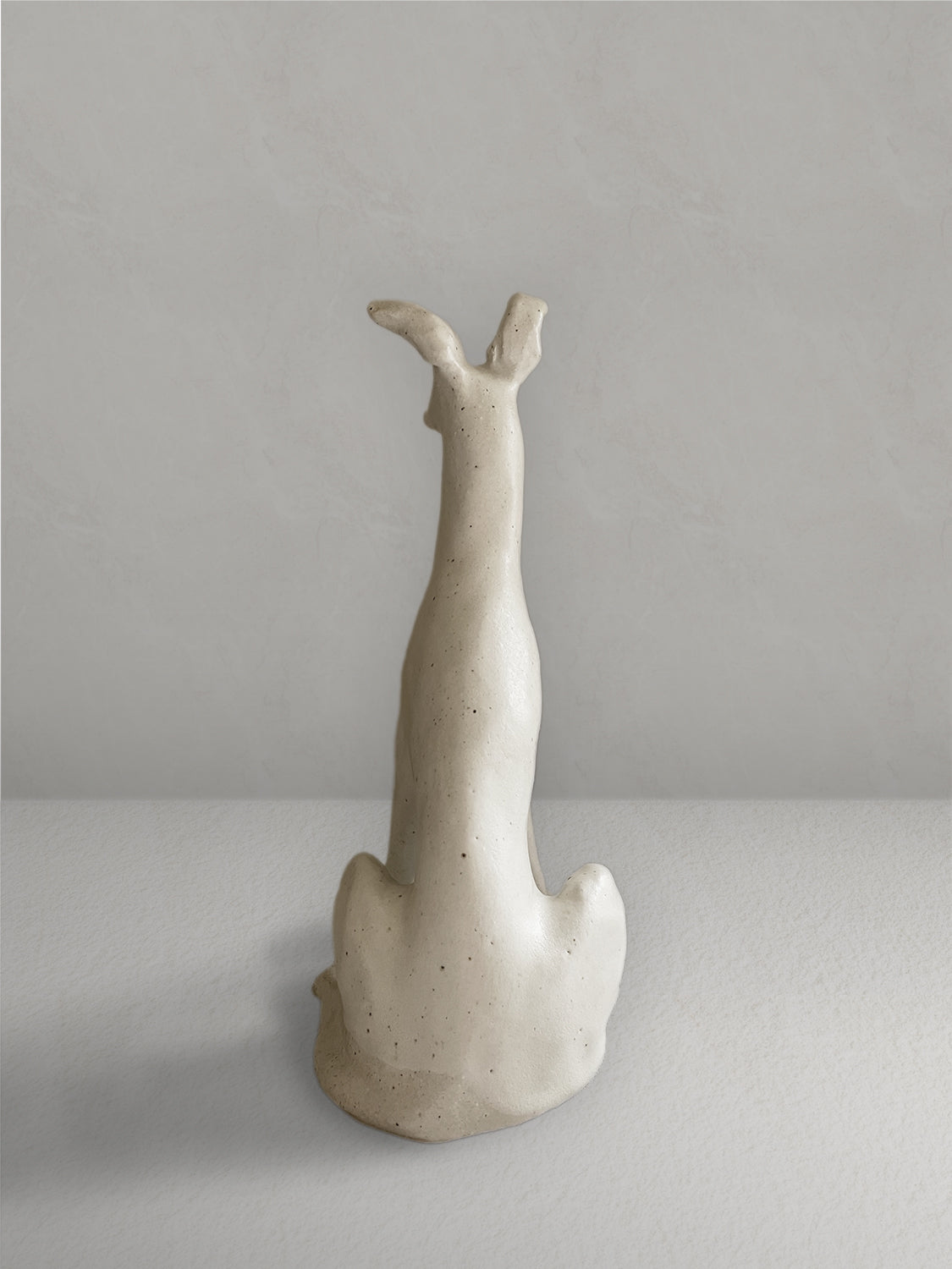 Seated stoneware sighthound sculpture with an off-white, lightly speckled glaze, on a light taupe surface with a textured taupe wall behind - back view.