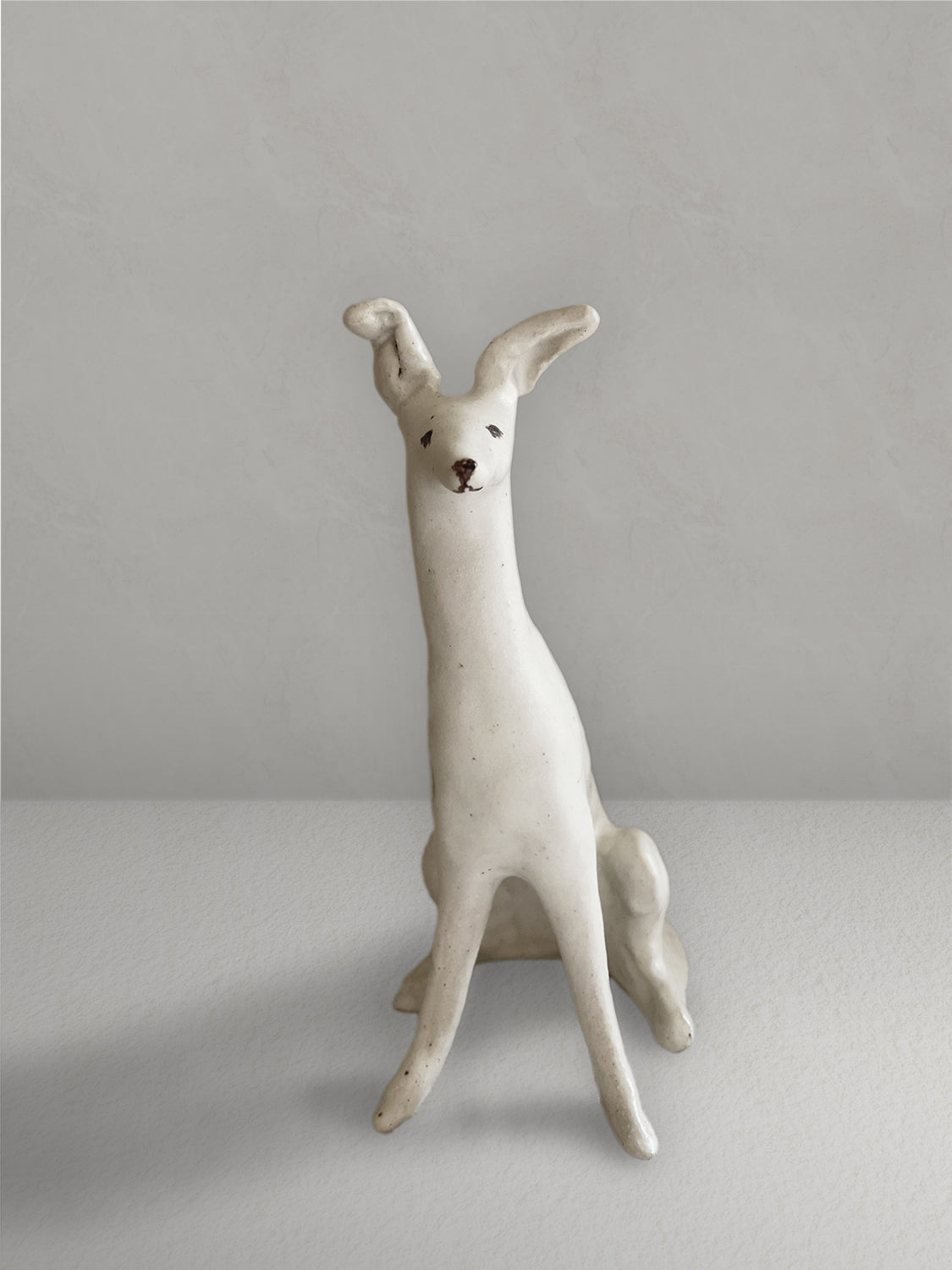 Seated stoneware sighthound sculpture with an off-white, lightly speckled glaze, on a light taupe surface with a textured taupe wall behind - front view.