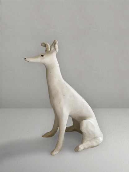 Seated stoneware sighthound sculpture with an off-white, lightly speckled glaze, on a light taupe surface with a textured taupe wall behind - left side view.