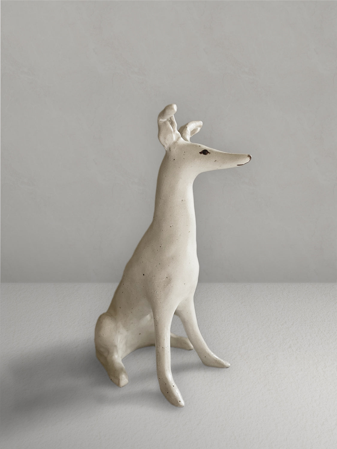 Seated stoneware sighthound sculpture with an off-white, lightly speckled glaze, on a light taupe surface with a textured taupe wall behind - right side view.