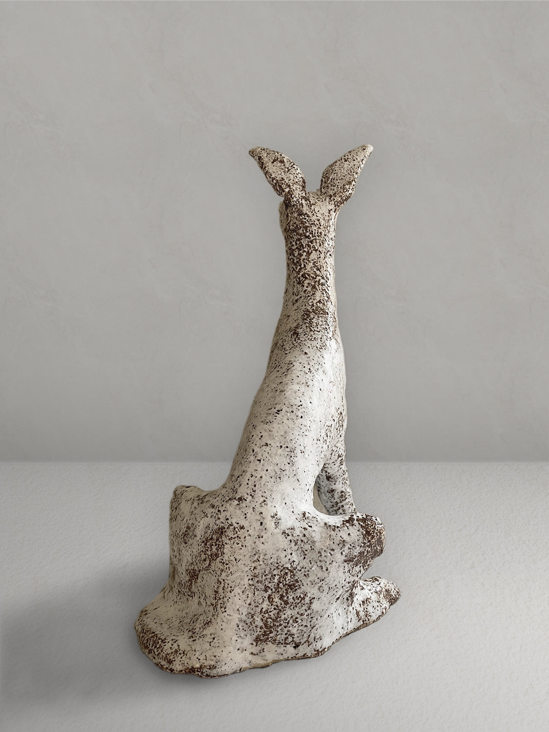 Seated stoneware sighthound sculpture with a textured, earthy red glaze, on a light taupe surface with a textured taupe wall behind - back view.