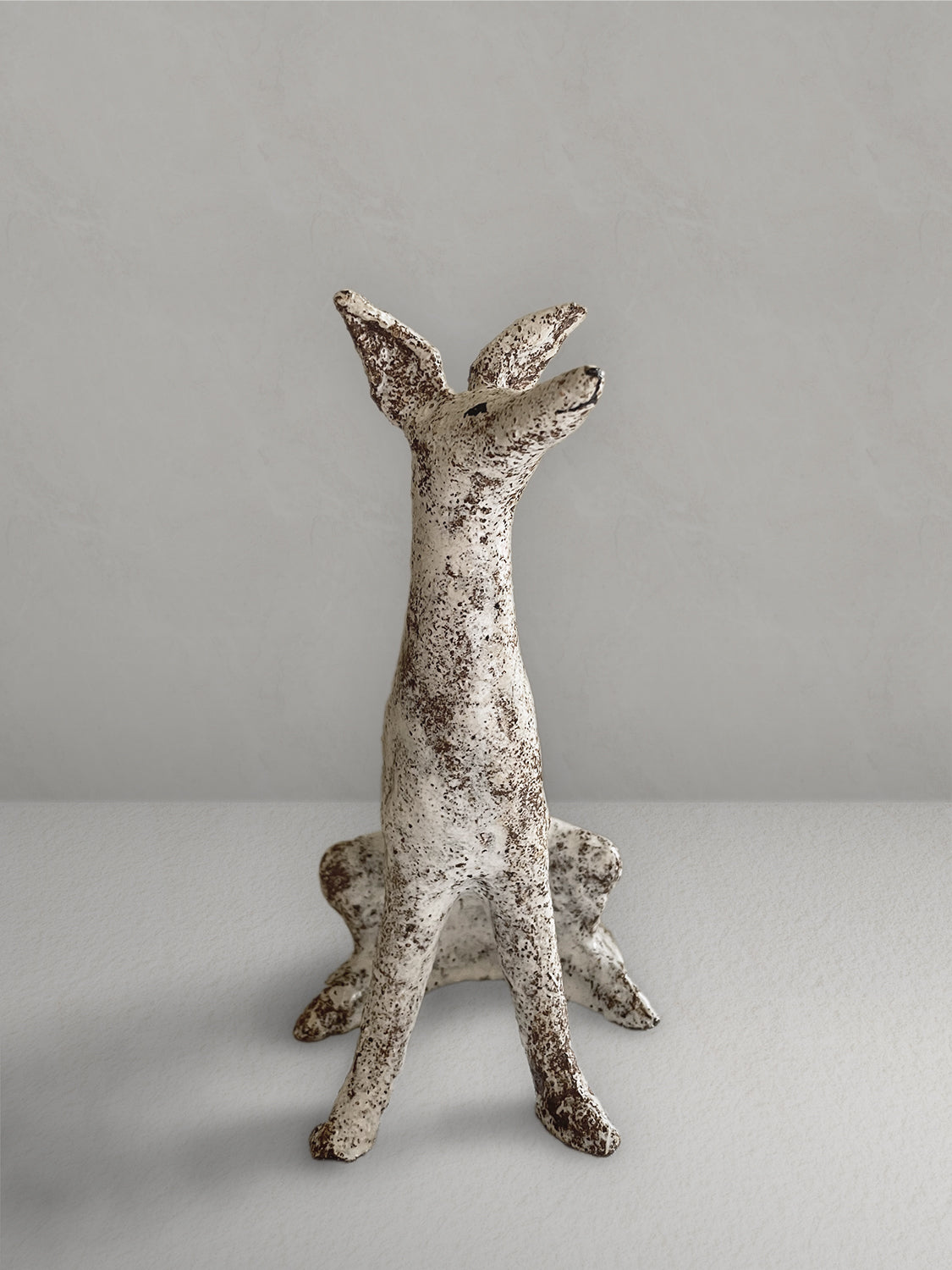 Seated stoneware sighthound sculpture with a textured, earthy red glaze, on a light taupe surface with a textured taupe wall behind - front view.