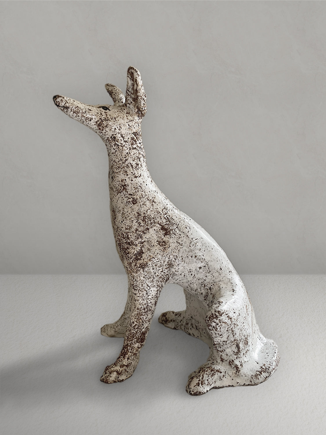 Seated stoneware sighthound sculpture with a textured, earthy red glaze, on a light taupe surface with a textured taupe wall behind - left side view.