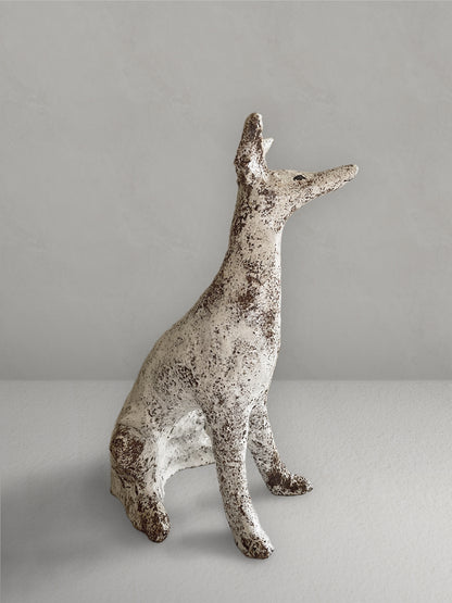 Seated stoneware sighthound sculpture with a textured, earthy red glaze, on a light taupe surface with a textured taupe wall behind - right side view.