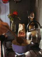 Person stirring a cup of hot chocolate with a spoon on a stove, with a bag of hot chocolate mix and a teapot in the background.