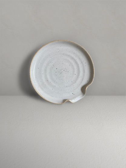 Ceramic spoon rest in glossy off-white speckled glaze on a light taupe surface with a textured taupe wall behind - front view.