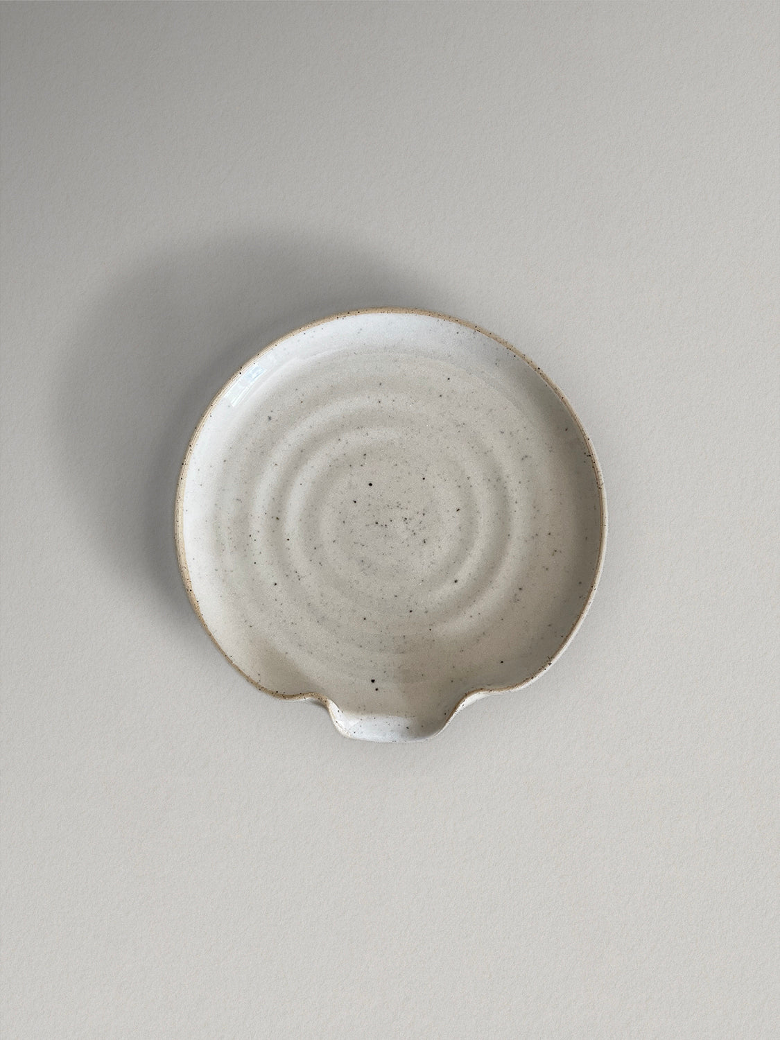 Ceramic spoon rest in glossy off-white speckled glaze on a light taupe surface - top view.