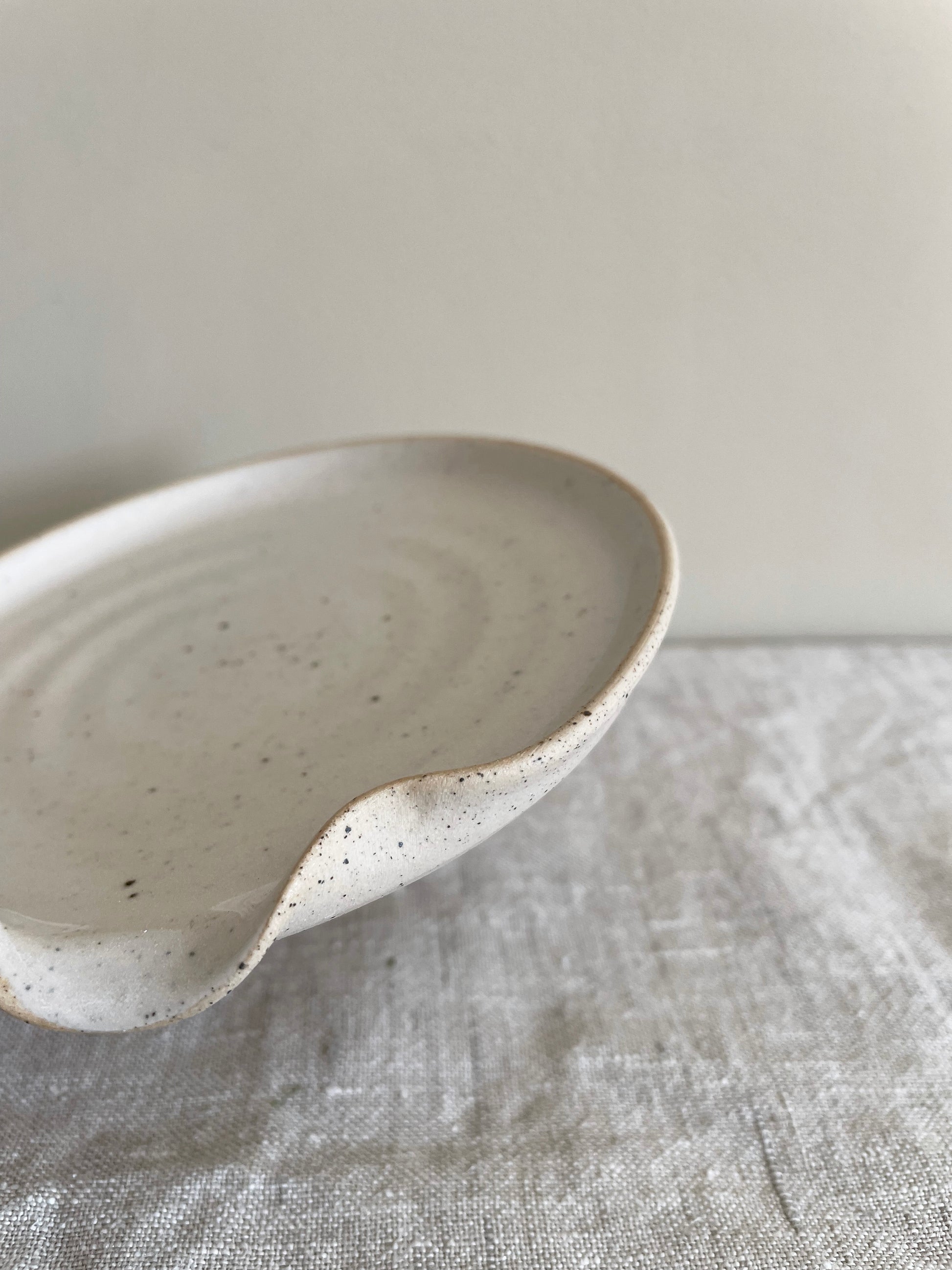 Ceramic spoon rest in glossy off-white speckled glaze on a beige linen surface with a beige painted wall behind - close-up view.