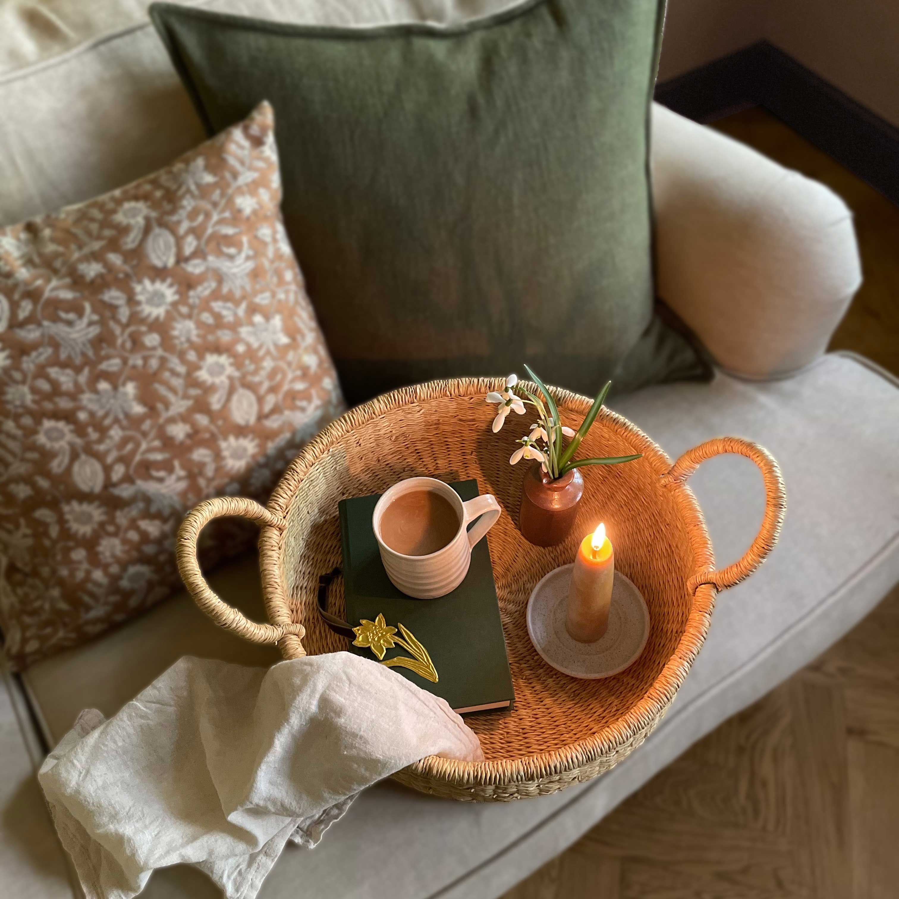 Handwoven round tray with a mug, book, candle and flowers on a linen sofa with cushions in the background.