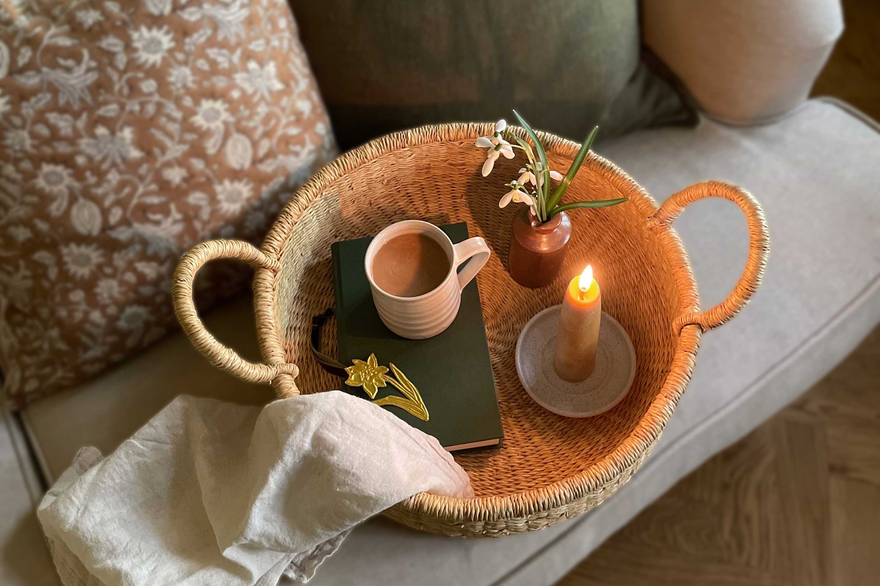 Handwoven round tray with a mug, book, candle and flowers on a linen sofa with cushions in the background.