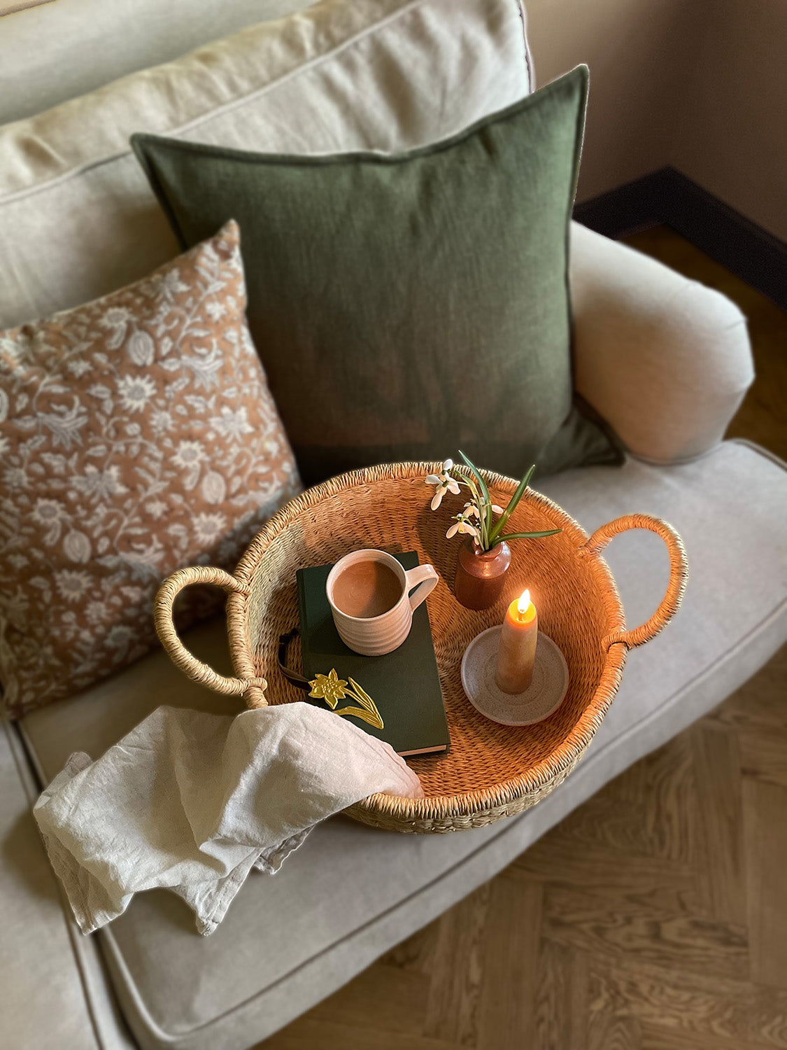 Handwoven round tray with a mug, book, candle and flowers on a linen sofa with cushions in the background.