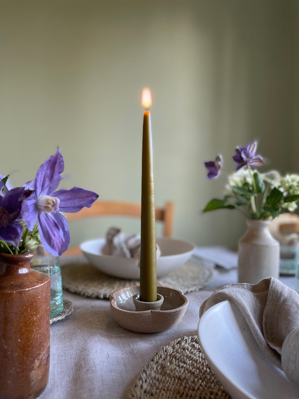 Taper candle in a ceramic holder on a dining table with flowers and dishes.