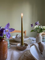 Taper candle in a ceramic holder on a dining table with flowers and dishes.