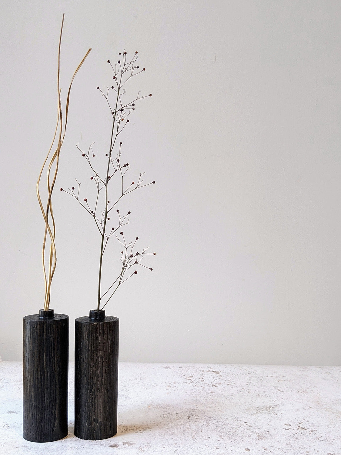 Pair of straight bud vases in ebonised Oak with dried stems, on a mottled off-white surface against a plain taupe wall - front view.