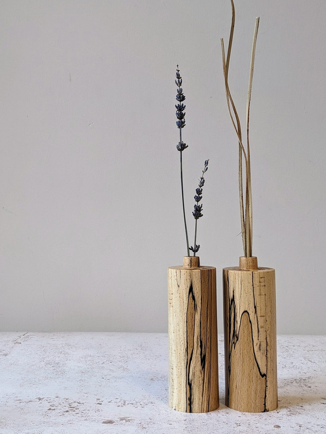 Pair of straight bud vases in spalted Beech with dried stems, on a mottled off-white surface against a plain taupe wall - front view.