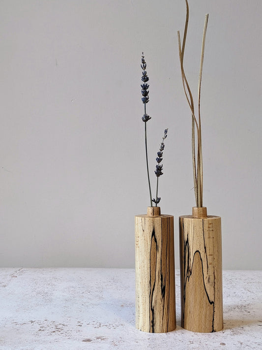 Pair of straight bud vases in spalted Beech with dried stems, on a mottled off-white surface against a plain taupe wall - front view.