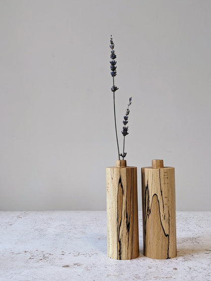 Pair of straight bud vases in spalted Beech with dried stems, on a mottled off-white surface against a plain taupe wall - front view.