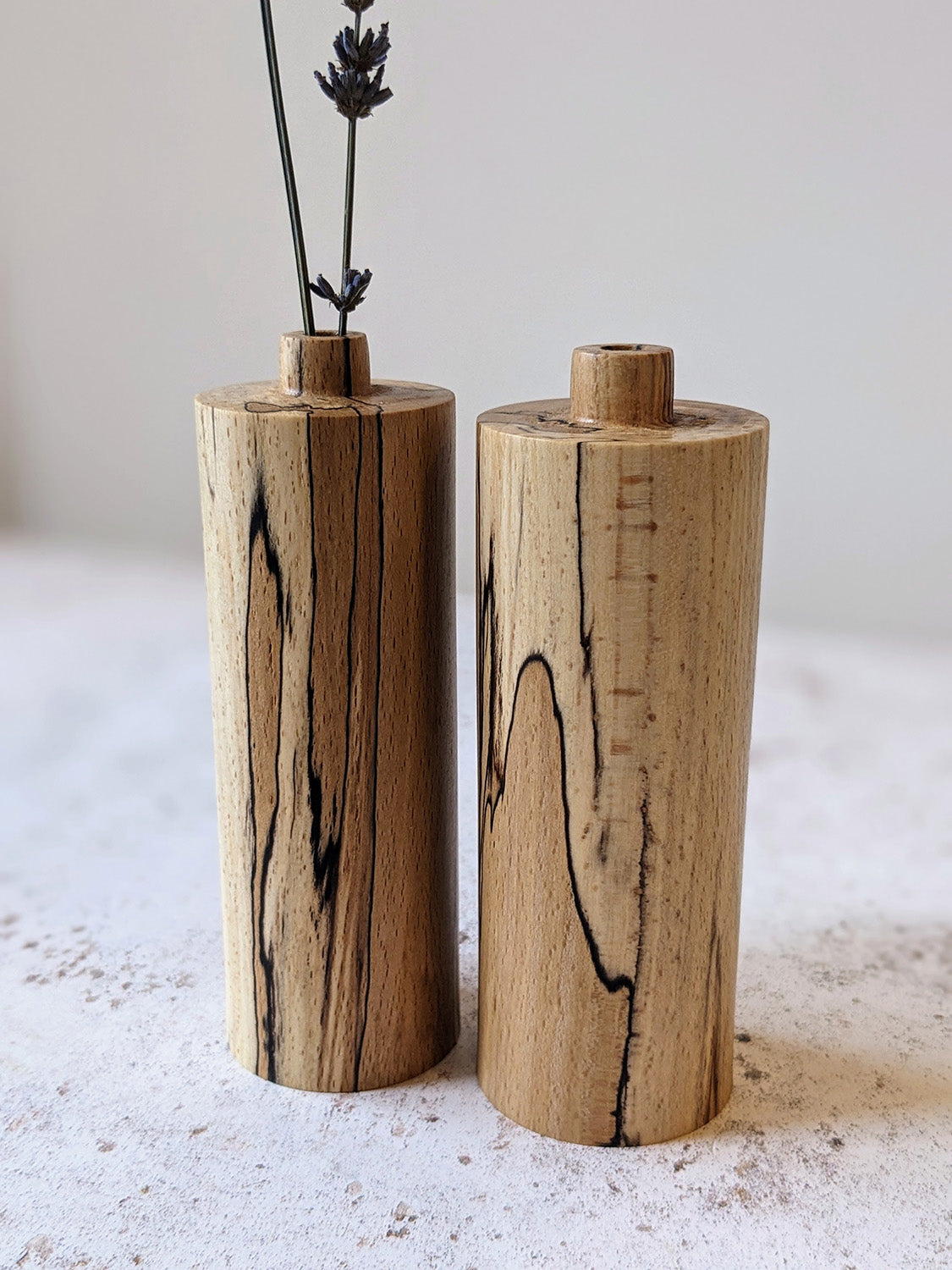 Pair of straight bud vases in spalted Beech with dried stems, on a mottled off-white surface against a plain taupe wall - front view close-up.