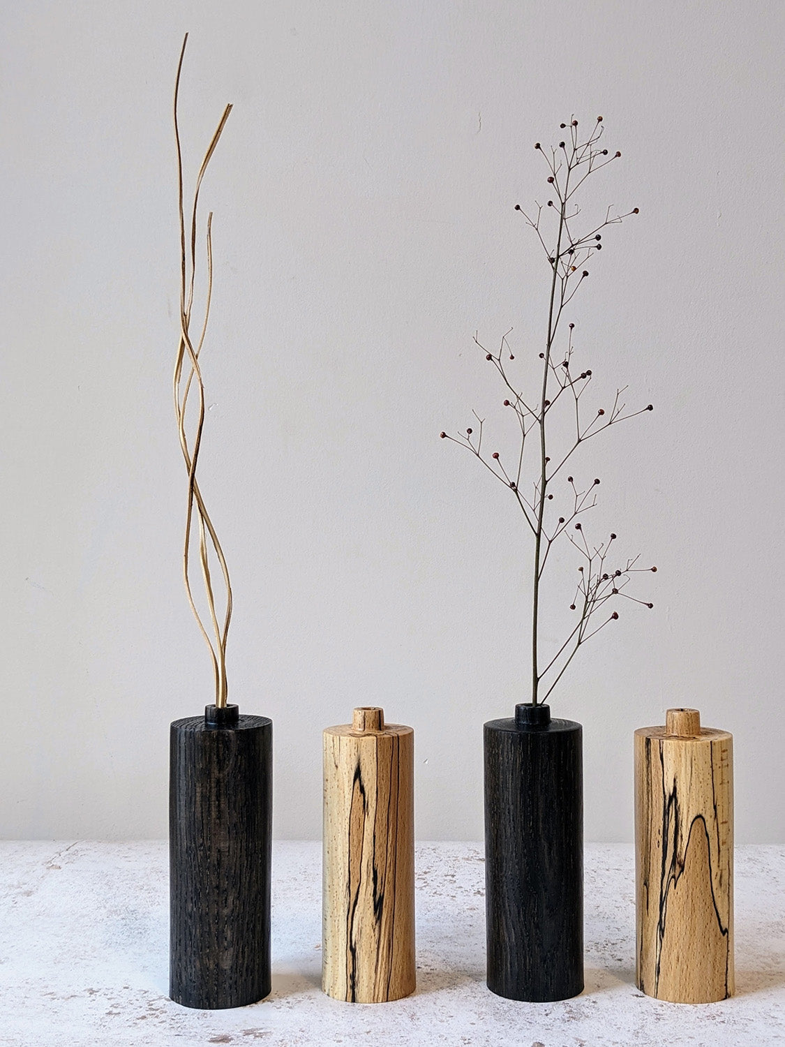 Two pairs of straight bud vases in ebonised Oak and spalted Beech with dried stems, on a mottled off-white surface against a plain taupe wall - front view.