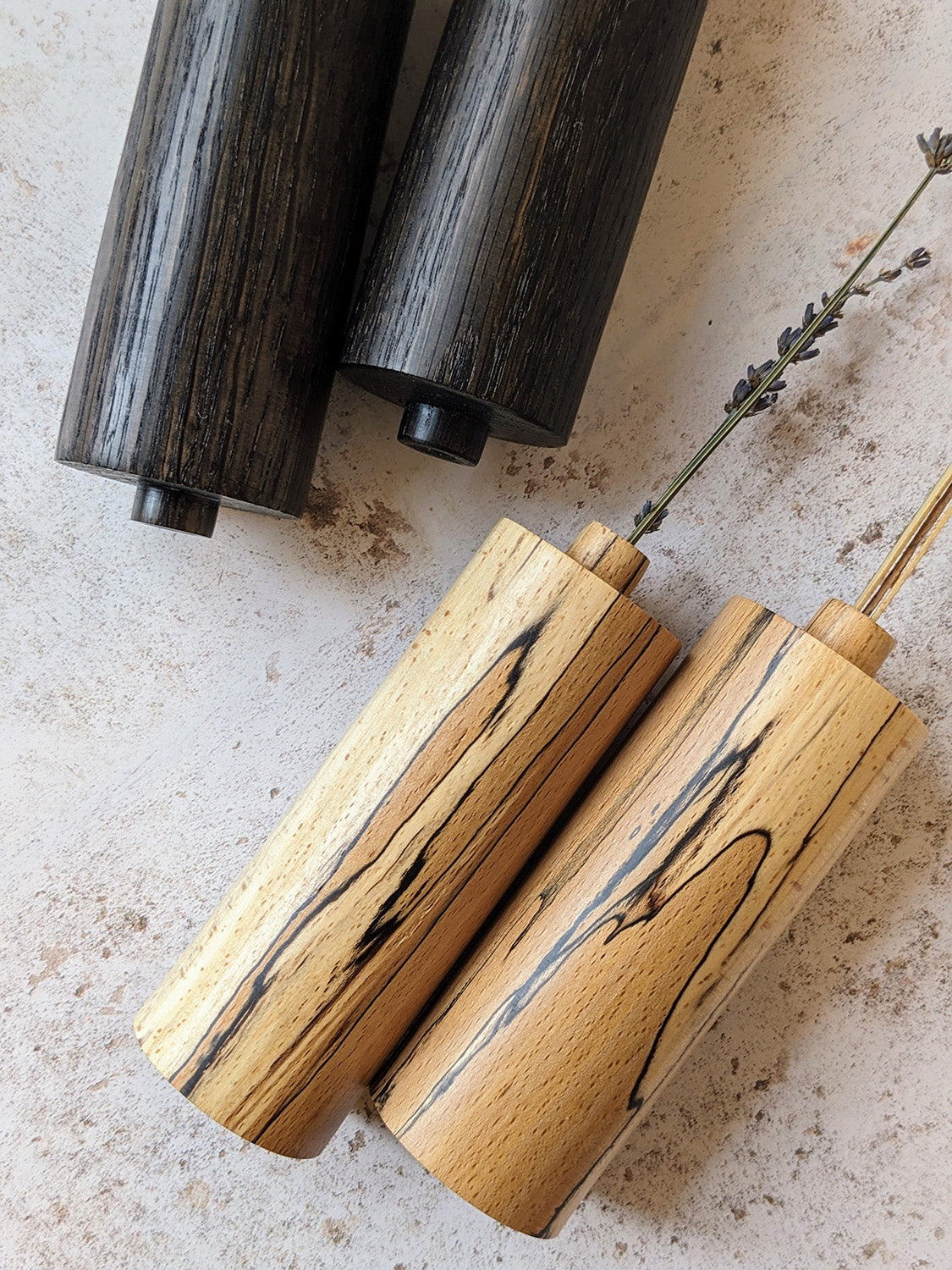 Two pairs of straight bud vases in ebonised Oak and spalted Beech with dried stems, on a mottled off-white surface - top view close-up.