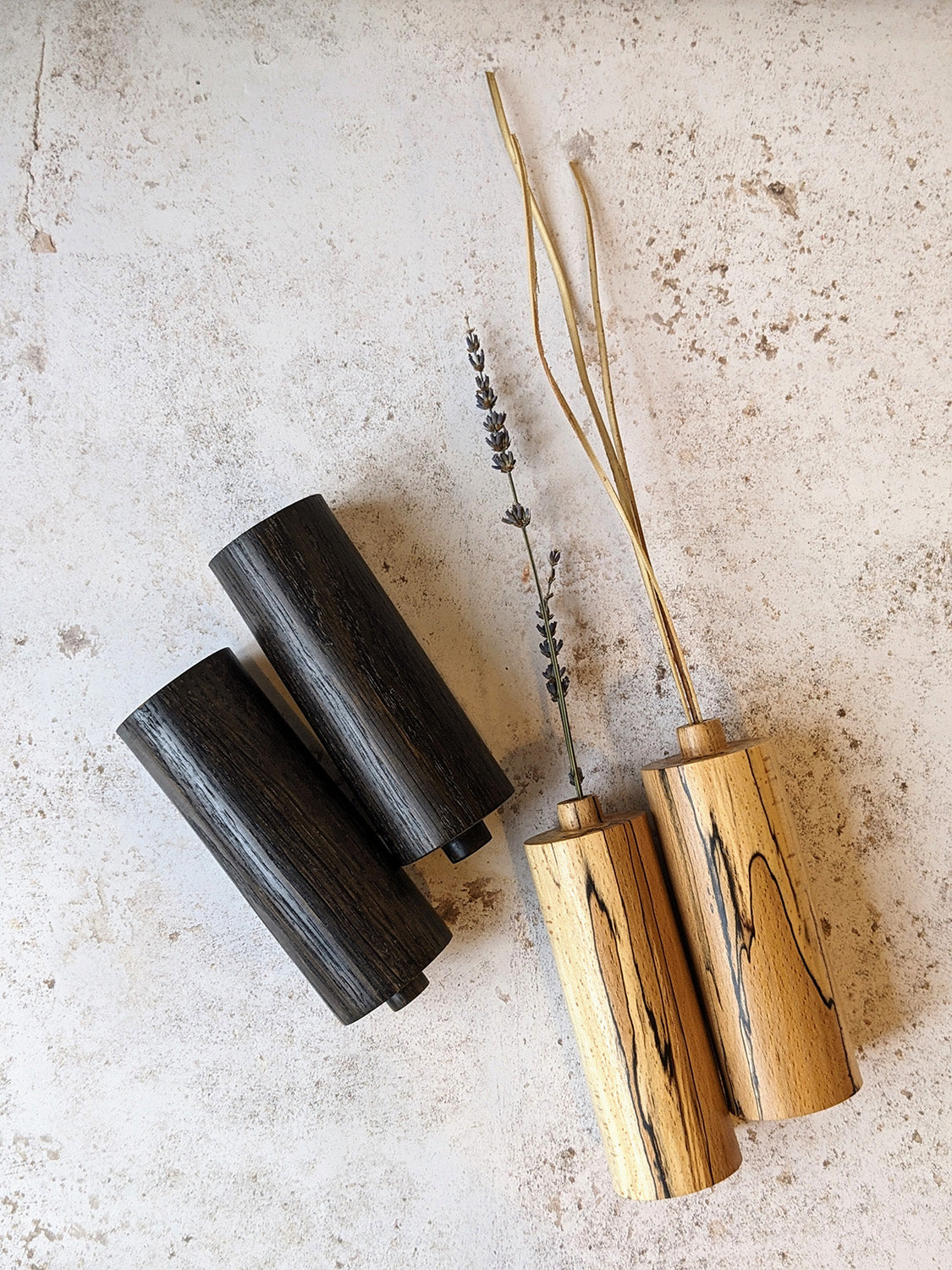 Two pairs of straight bud vases in ebonised Oak and spalted Beech with dried stems, on a mottled off-white surface - top view.