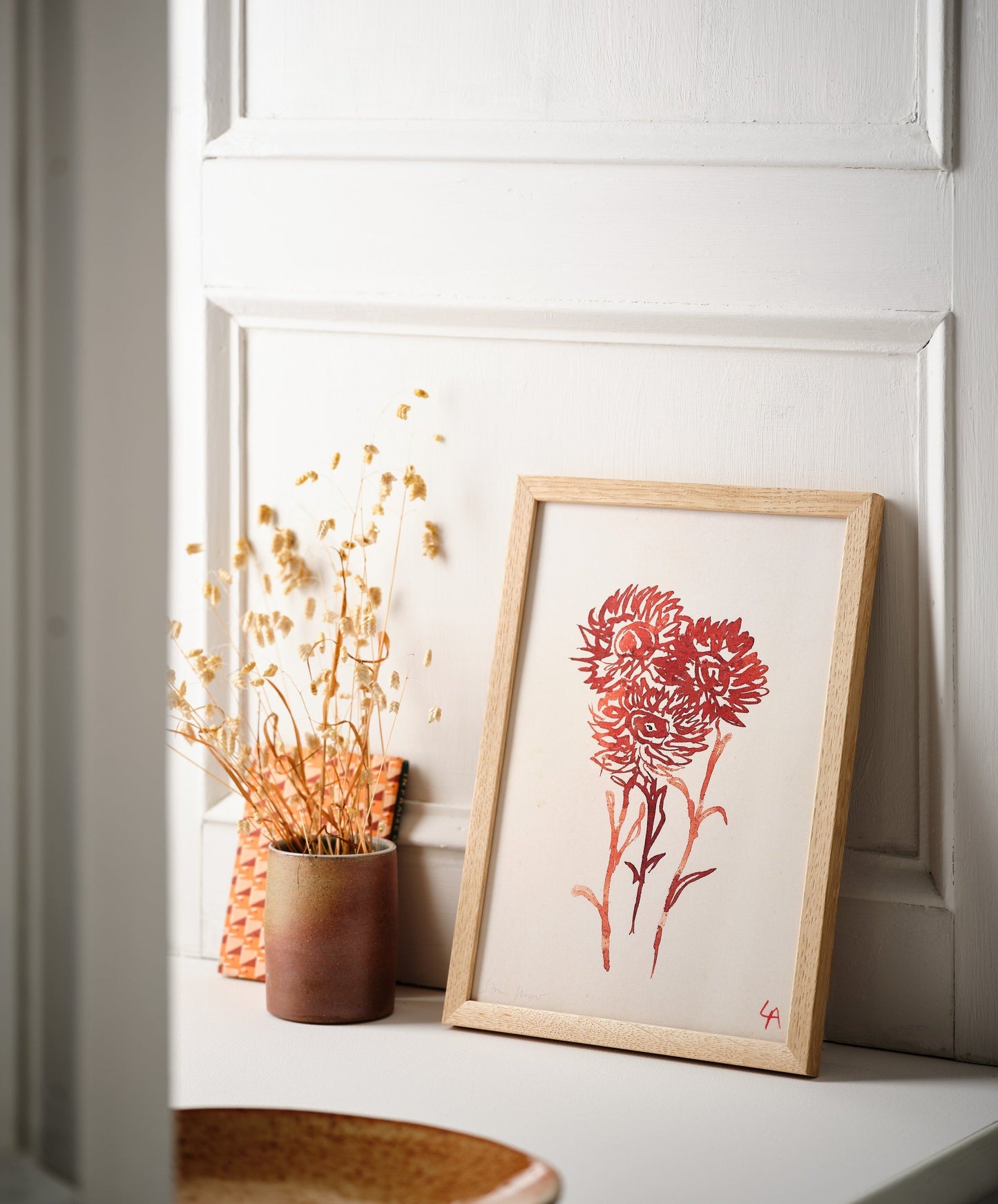 Art print of three Strawflowers in red ink on vintage paper in a natural oak frame, sitting on a white painted windowsill alongside a stoneware vessel with dried flowers