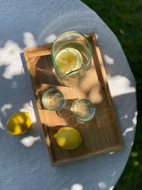 Two handmade recycled glass tumblers and a carafe with lemons on a wooden tray outdoors.