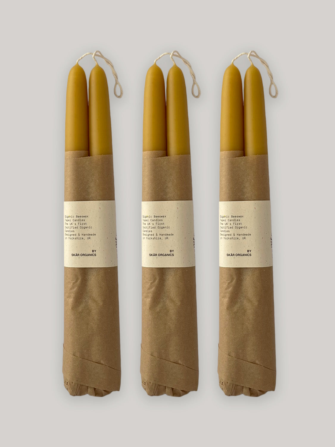 Three pairs of beeswax taper candles wrapped in brown paper with labels on a neutral background.