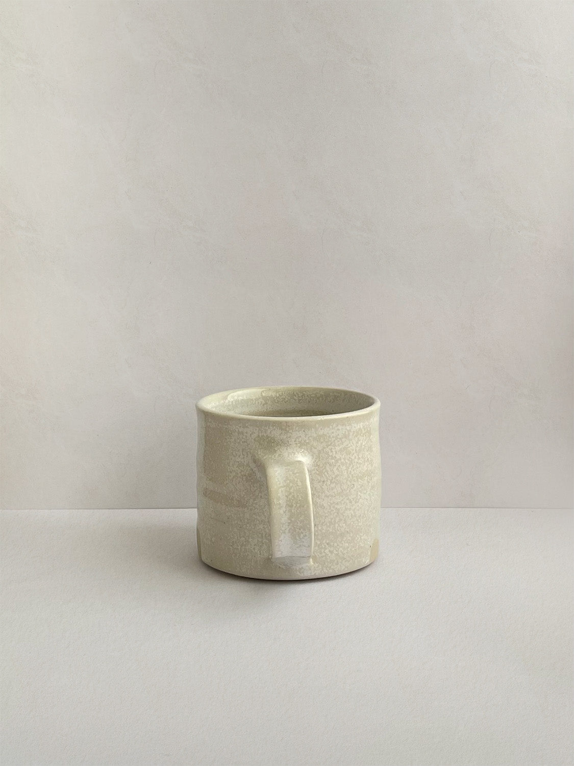 A beige handmade ceramic espresso mug with a handle, on a neutral background - back view.