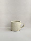 A beige handmade ceramic espresso mug with a handle, on a neutral background - left side view.