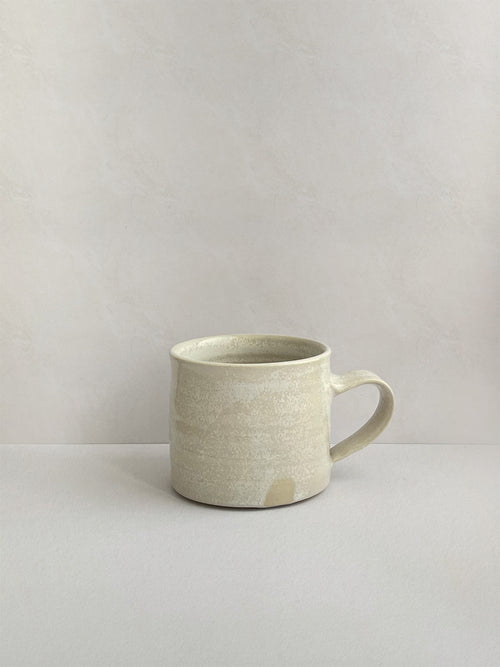 A beige handmade ceramic espresso mug with a handle, on a neutral background - left side view.